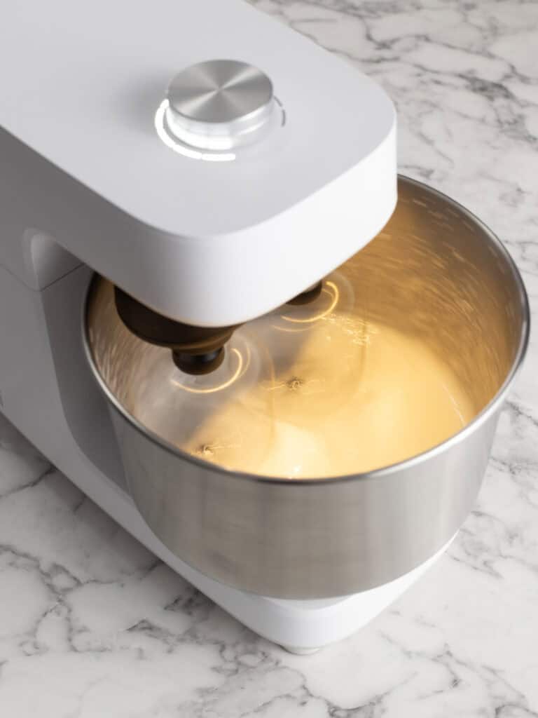 A white stand mixer with a stainless steel bowl containing a light-colored mixture being whipped by the beaters.