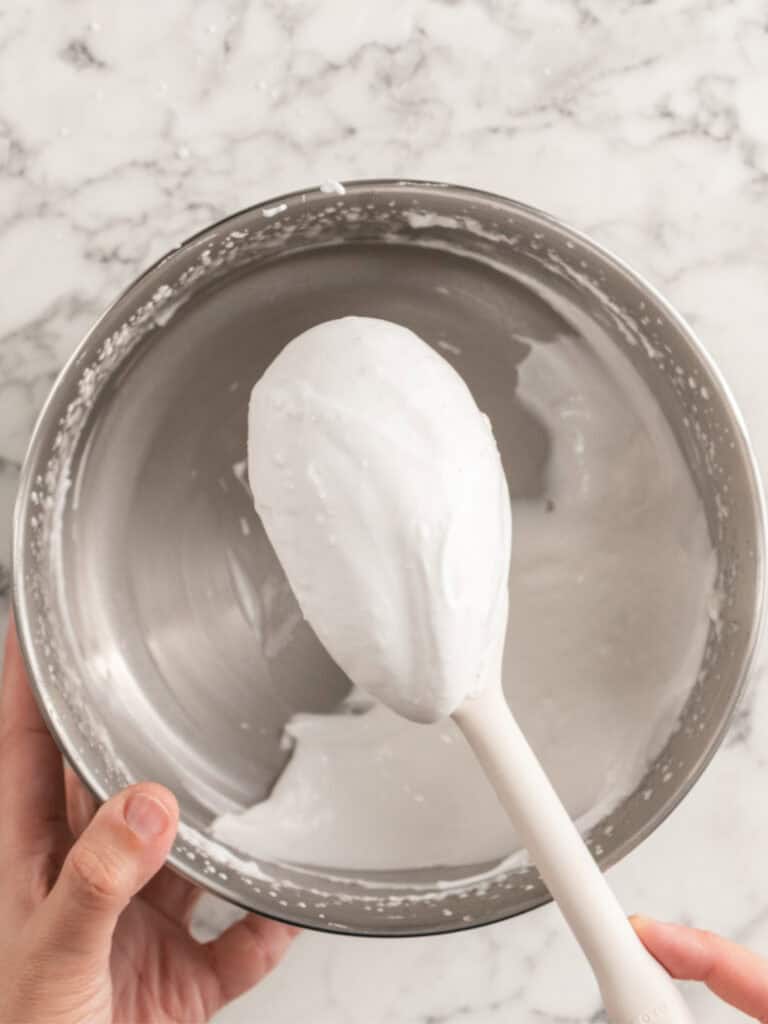 A spatula covered in white whipped meringue held over a metal mixing bowl.