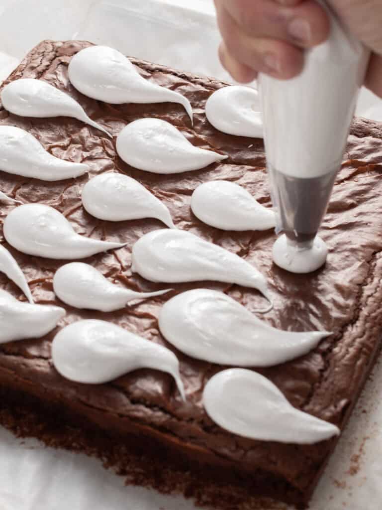 A hand uses a piping bag to add white icing droplets to the top of a square brownie.
