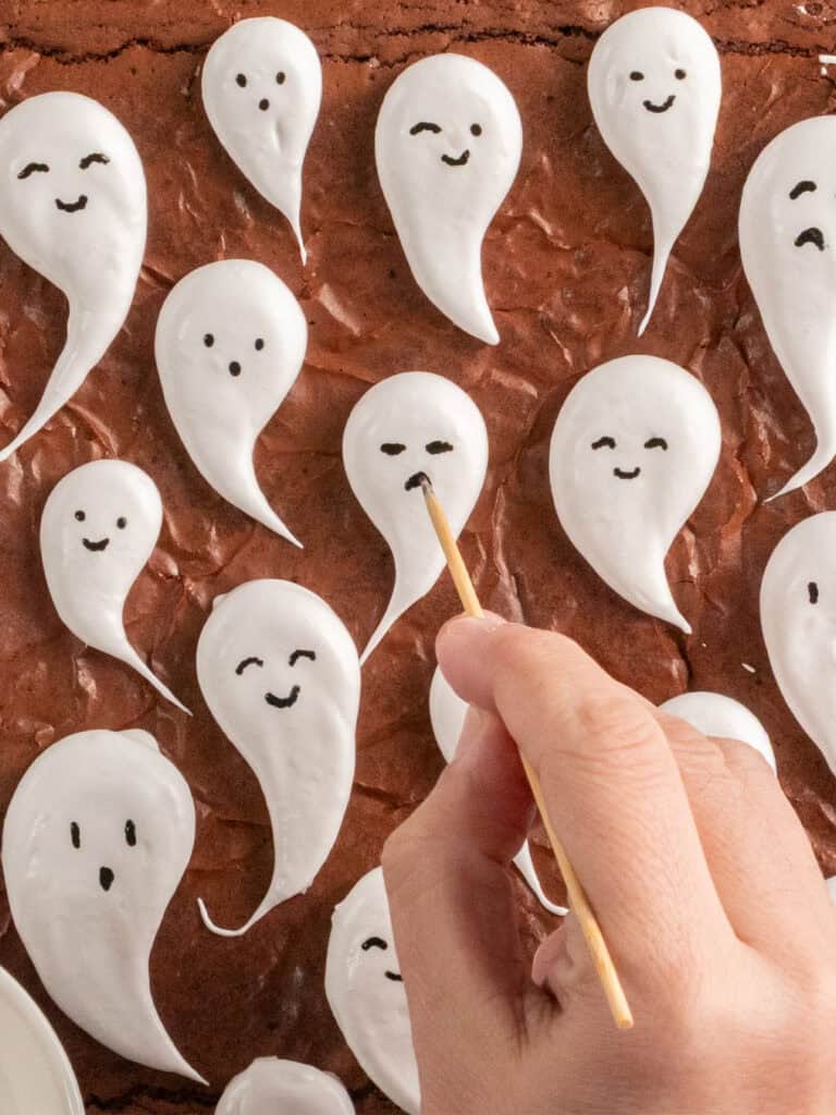 A hand uses a wooden toothpick to draw a mouth on a white icing ghost atop a chocolate brownie.
