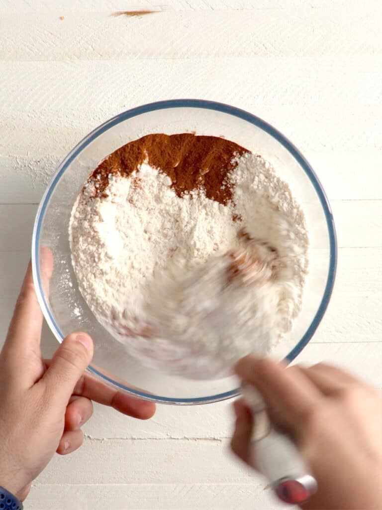 Hands mixing flour and cocoa powder in a glass bowl.
