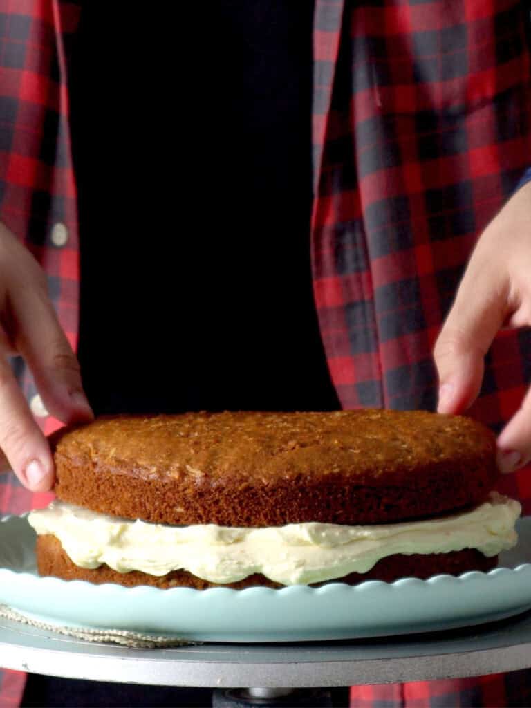 Hands placing a top cake layer onto a base layer filled with white frosting on a light blue scalloped plate.