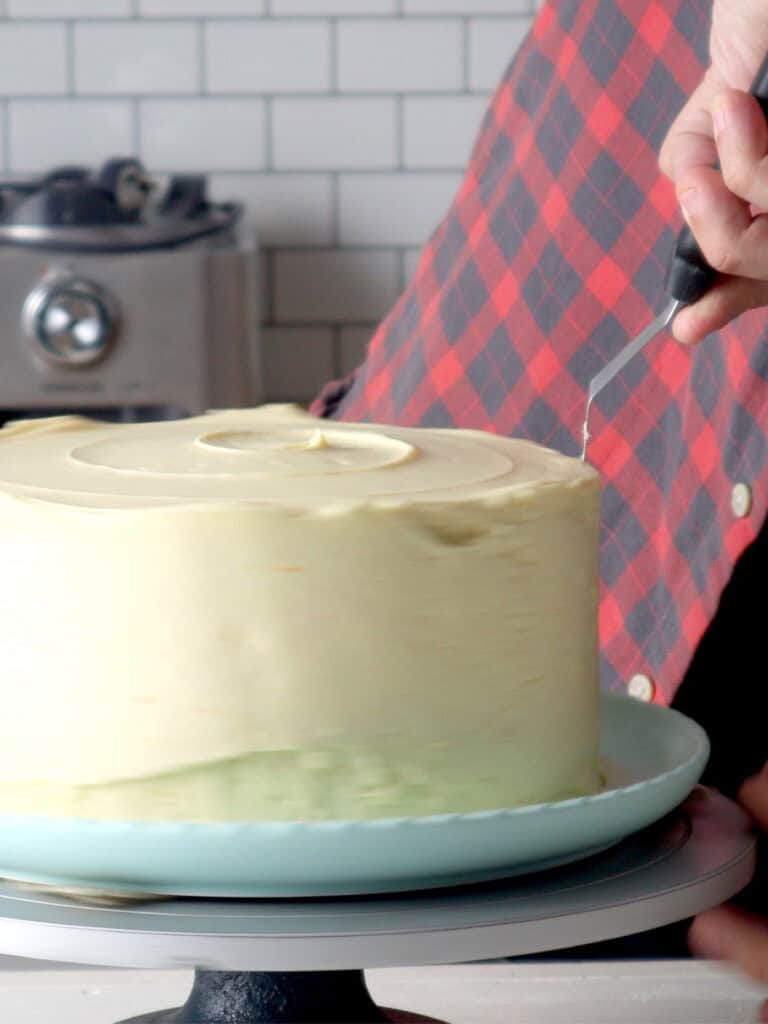 A person in a red and black plaid shirt uses an offset spatula to smooth the frosting on a round cake.