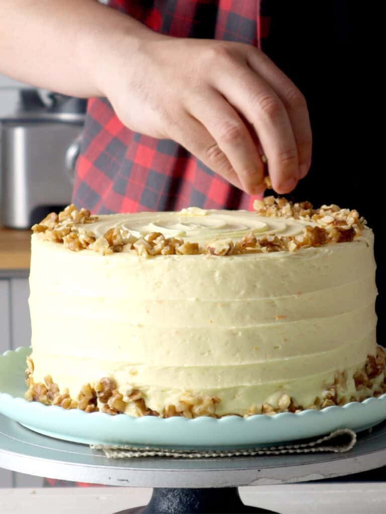 A persons hand sprinkles chopped walnuts onto the top of a frosted layer cake on a pedestal stand.