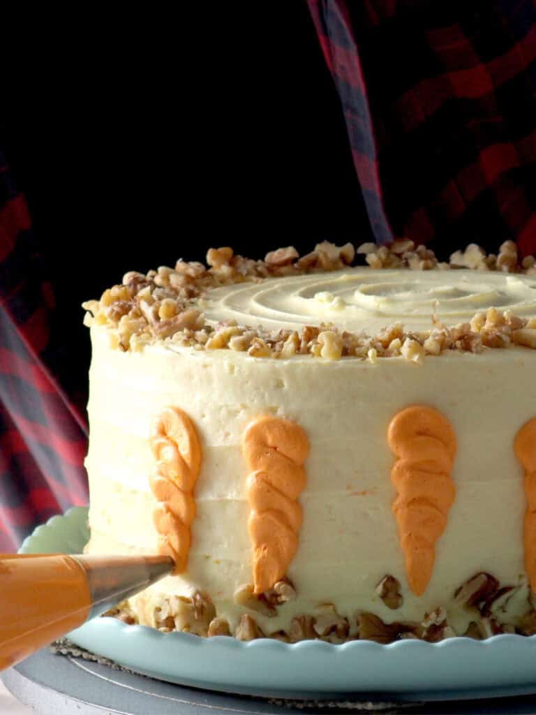 A frosted carrot cake being decorated with orange icing carrots, topped with chopped walnuts on a light blue cake stand.