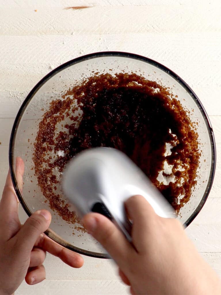 A hand uses an electric hand mixer to blend dark brown ingredients in a glass bowl.