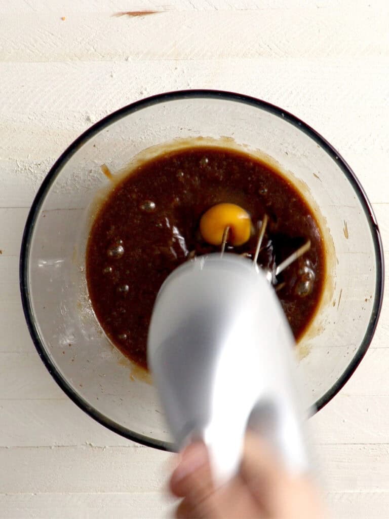 An electric hand mixer blends an egg into a dark brown mixture in a glass bowl.