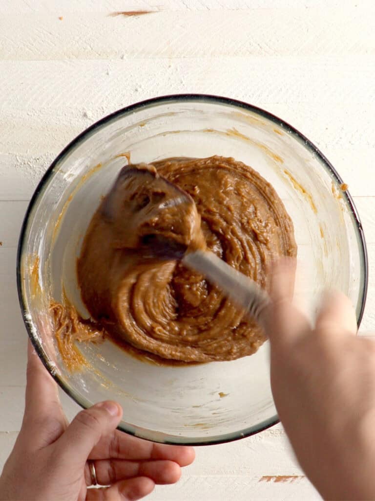 A person mixes brown cookie dough in a glass bowl with a spatula.