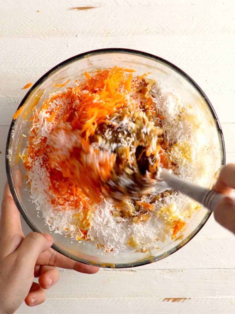 A hand stirs shredded carrots, shredded coconut, and chopped nuts in a glass mixing bowl.