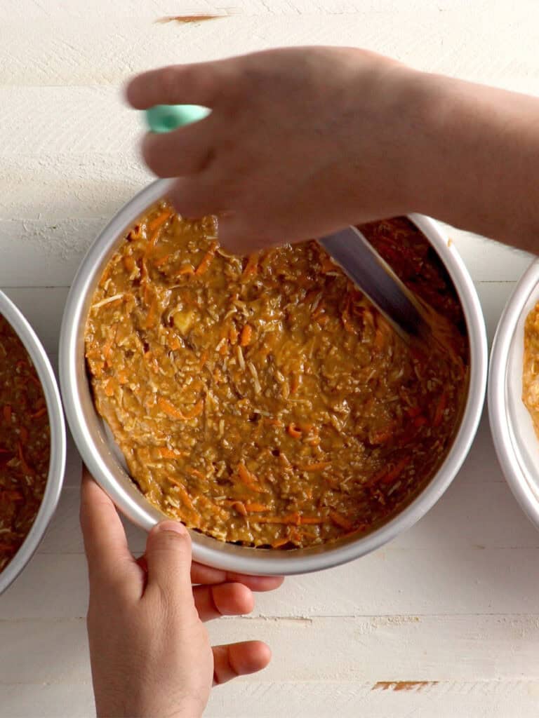 Hands spreading carrot cake batter into a round metal baking pan with a spatula.