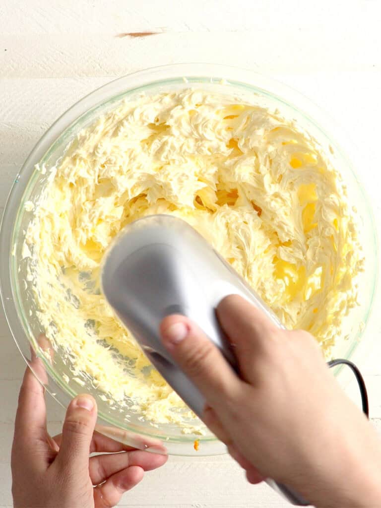 A hand uses a handheld electric mixer to whip yellow frosting in a glass bowl.