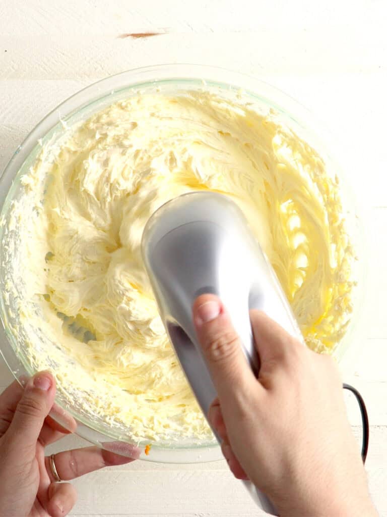 A hand uses an electric hand mixer to blend pale yellow frosting in a glass bowl.