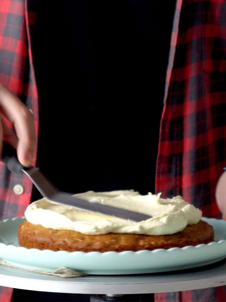 A person in a red plaid shirt uses an offset spatula to frost a cake on a light blue cake stand.